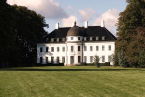 Et stort hvidt palæ med sort tag, centralt rundt tårn og flere skorstene, omgivet af træer og en stor, pænt klippet grøn græsplæne under en delvis overskyet himmel.