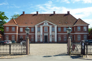 Et stort, historisk palæ i røde mursten med hvide søjler, symmetriske vinduer og rødt tegltag, set bag en smedejernsport på en solrig dag. Biler er parkeret på en grusbelagt indkørsel foran.