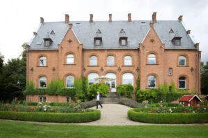 En person går på en grussti foran et stort palæ i røde mursten med et gråt skifertag, buede vinduer og efeu, der klatrer op ad væggene. Velplejede haver og grønne hække omgiver bygningen.