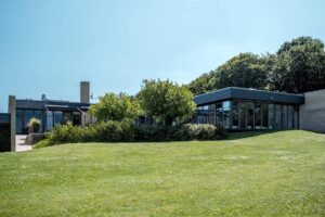En moderne bygning i én etage med store vinduer ligger på en grøn græsplæne omgivet af træer og buske under en klar blå himmel.