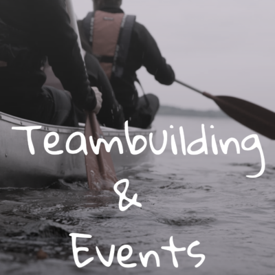 To personer padler i en kano på et roligt vand, iført redningsveste. Ordene Teambuilding & Events er skrevet på tværs af billedet med hvid tekst.