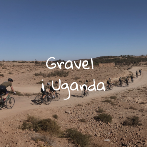 En gruppe cyklister kører ad en snoet grusvej gennem et tørt, stenet landskab under en klar blå himmel. Teksten Gravel i Uganda er skrevet hen over billedet.