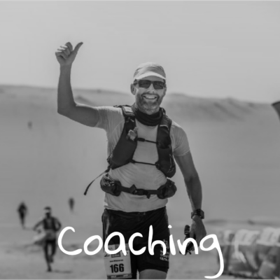 En smilende mand iført løbetøj og startnummer giver tommelfingeren op, mens han løber i et ørkenløb. Ordet Coaching er skrevet i bunden af billedet. Andre løbere er synlige i baggrunden.