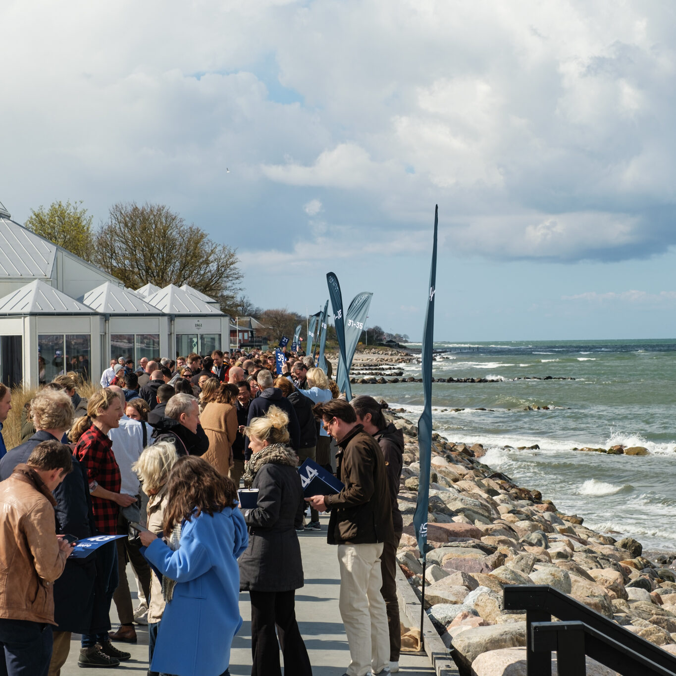 En stor gruppe mennesker er samlet på en asfalteret gangbro langs en klippekyst med bølger i nærheden af hvide, moderne bygninger under en delvis overskyet himmel. Nogle holder papirer i hånden eller chatter i små grupper.