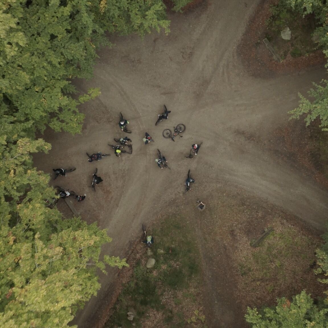 Luftfoto af en gruppe cyklister, der er samlet ved et jordkryds omgivet af tætte, grønne træer i en skov. Cyklisterne og deres cykler er spredt i krydset.