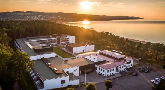 Luftfoto af et stort hvidt bygningskompleks omgivet af træer, beliggende nær en kystlinje ved solnedgang, med roligt hav og fjerne bakker under en delvist overskyet himmel.