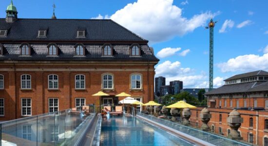 En pool på taget med glassider ligger ved siden af en historisk murstensbygning. Gule parasoller skygger for liggestole, og moderne bygninger og en høj forlystelsespark kan ses i baggrunden under en blå himmel med skyer.