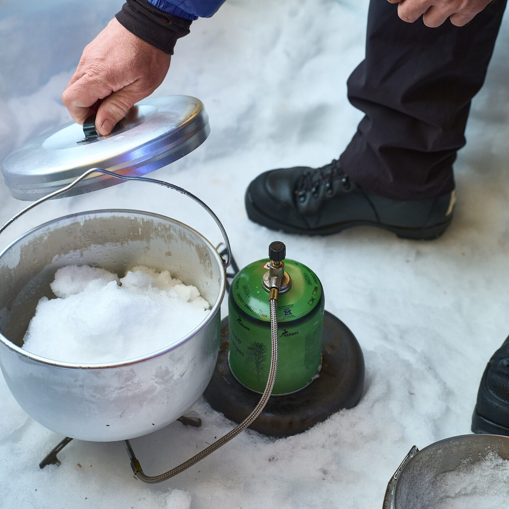 En person udendørs i sneen løfter låget af en metalgryde fyldt med sne, der er placeret på et grønt campingkomfur. Scenen viser vinterstøvler og sne på jorden.