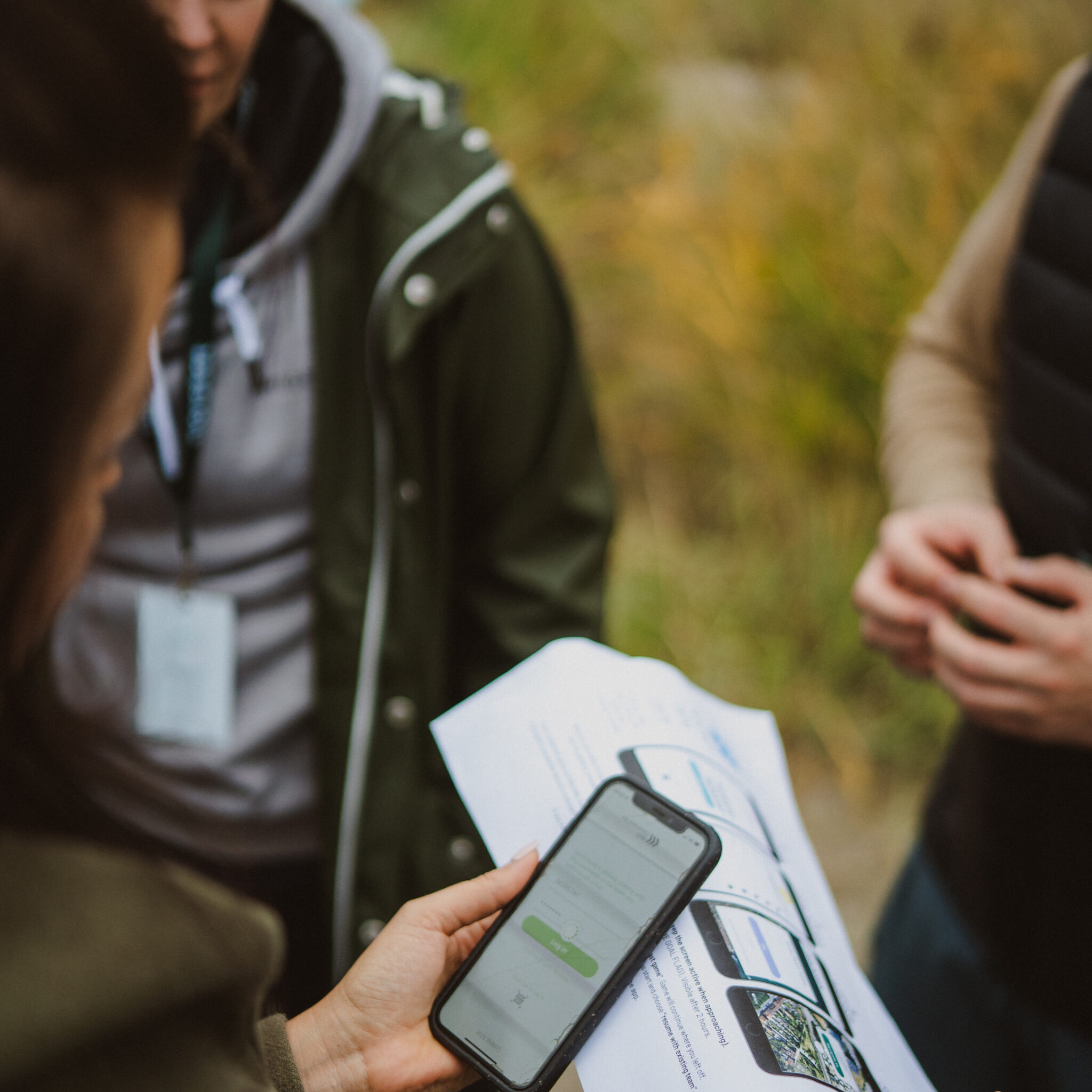 To personer står udendørs og kigger på et trykt dokument med billeder, mens den ene holder en smartphone hen over papiret. Begge har jakker på, og baggrunden er sløret grønt.