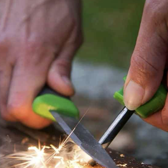 Nærbillede af hænder, der bruger en kniv med grønt skaft og en ildstarter til at skabe gnister over et stykke træ udendørs.