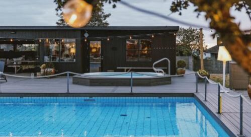 En moderne udendørs pool med klart blåt vand er i forgrunden, mens et spabad står på et trædæk foran et moderne, mørkfarvet hus. Lyskæder og træer skaber en hyggelig atmosfære.