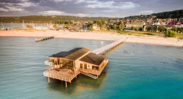 En træbygning på pæle strækker sig ud over klart blåt vand og er forbundet med en sandstrand via en lang mole. I baggrunden ses en kystby og grønne bakker under en delvis overskyet himmel.
