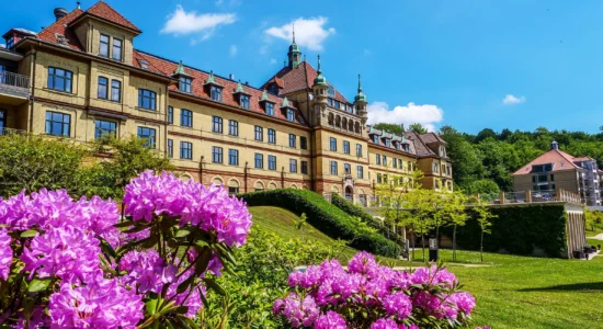 En stor, historisk bygning med gule murstensvægge og røde tegltage står bag blomstrende lyserøde blomster og grønne plæner under en lyseblå himmel. I baggrunden ses træer og en anden bygning.
