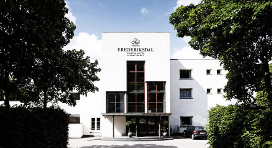 Hvid, moderne bygning med store vinduer og Frederiksdal Sinatur Hotel & Konference-skilt over indgangen; træer på begge sider indrammer hotellet, og en bil er parkeret delvist synligt til højre.