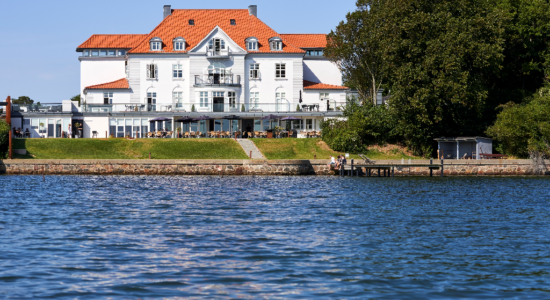 En stor hvid bygning med rødt tegltag ligger ved vandkanten, omgivet af træer. Der er en bådebro, et udendørs siddeområde og en stensti, der fører ned til vandkanten.